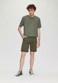 Zielona bawełniana koszulka w kolorze oliwkowym w połączeniu z oliwkowymi szortami. Szorty mają podwinięte nogawki. Model nosi oliwkowe trampki za kostkę z białymi sznurówkami.