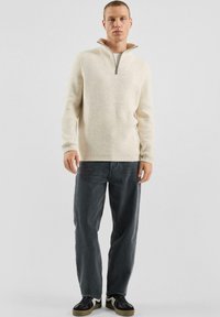 Roomkleurige geribbelde gebreide pullover met een hals met kwartritssluiting, gecombineerd met donkergrijze rechte jeans en zwarte sneakers met witte accenten.