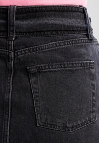 Detailní záběr na zadní kapsu a páskovou část černých džín s viditelnými smyčkami na pásek, které jsou nošeny s růžovou košilí zasunutou dovnitř.
