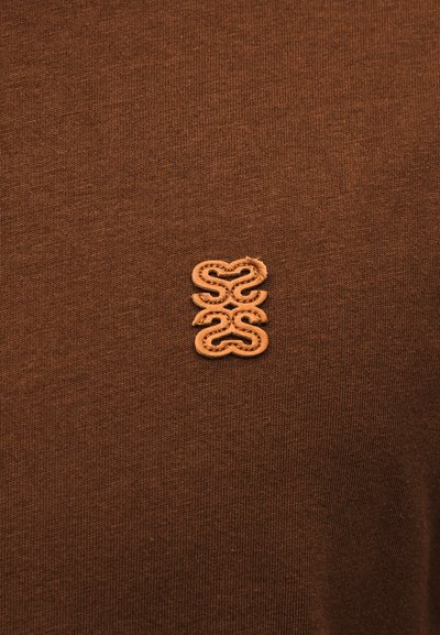 Tissu marron avec une petite pièce de broderie en relief présentant un motif symétrique et abstrait en fil marron plus clair.
