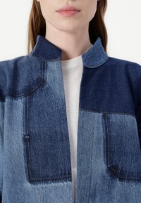 Τζάκετ από τζιν σε διάφορες αποχρώσεις του μπλε, με γιακά, και σχέδιο patchwork που διαθέτει αντιθετικά υφάσματα και ορατές ραφές.
