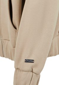 Beige hoodie med en blød tekstur, ribbede ærmer og en lille sort logoaccent på ærmet. Inkluderer et kængurulomme-design.