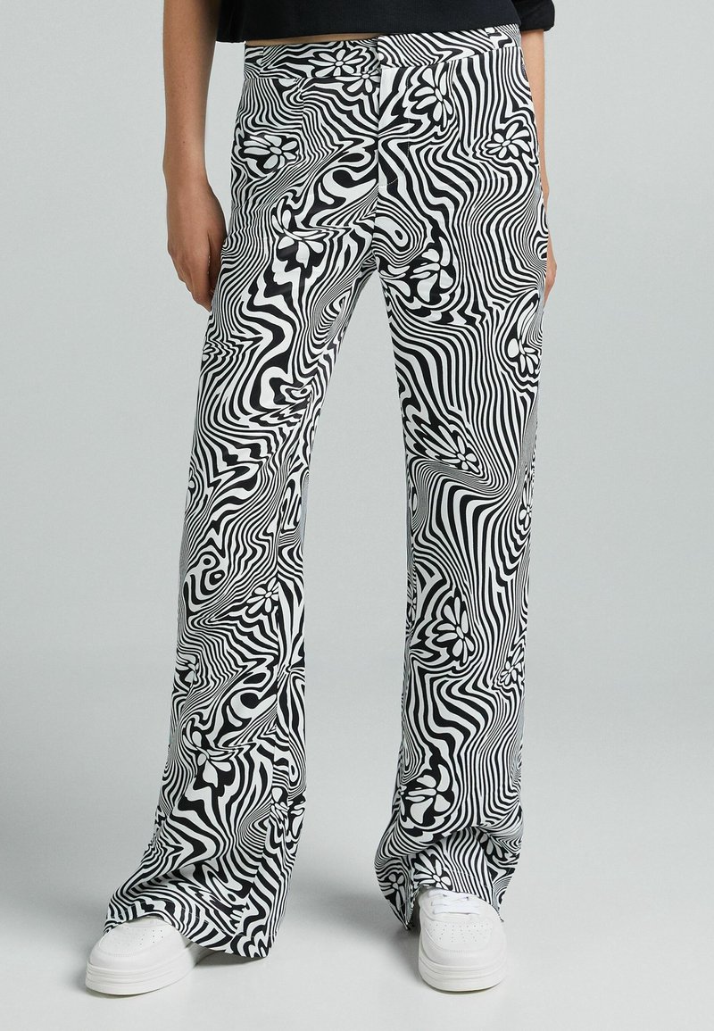 Svartvit mönstrade byxor med en vågig design och blommiga detaljer, tillverkade av ett mjukt tyg, kombinerat med vita sneakers.