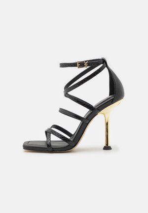 Sandale noire à talons hauts avec brides, bout carré, talon stiletto doré et bride de cheville avec boucle dorée et logo MK.
