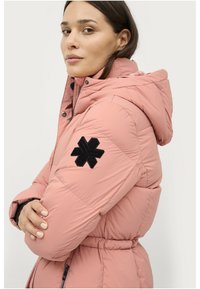 Lichtroze pufferjack met capuchon, voorzien van een zwarte sneeuwvlokpatch op de linker mouw en een cinched taille. Gelaagd stof met een gequilt ontwerp.