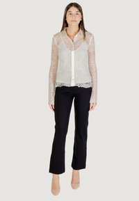 Camicia in pizzo a maniche lunghe con colletto, color crema, con motivo floreale trasparente e orlo a scallop, abbinata a pantaloni neri aderenti.