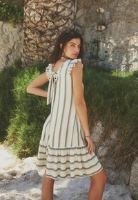 Žena stojící venku vedle kamenné zdi a zeleně, oblečená ve slonovinově bílých šatech bez rukávů se svislými a vodorovnými šedými pruhy a mašlí na zádech.