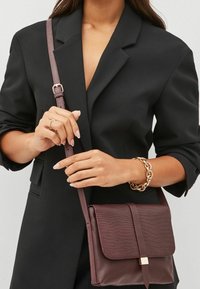 Sac bandoulière bordeaux avec un motif crocodile texturé, accents en métal doré, et une sangle mince et réglable. Porté avec un blazer noir.
