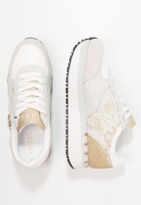 Hvide og gyldne atletik sneakers med snøredetaljer, teksturerede materialer, polstret krave og en tyk sål med gyldne accenter og nitter.