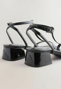 Sandalias de charol negro con tiras cruzadas, hebilla ajustable en el tobillo y tacones geométricos en bloque. Superficie lisa, diseño minimalista.