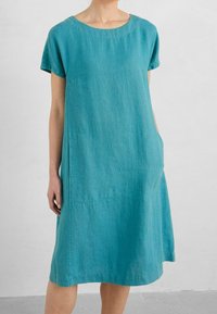 Ein locker sitzendes türkisfarbenes Kleid aus leichtem Stoff, mit kurzen Ärmeln, einem runden Ausschnitt und seitlichen Taschen mit geschwungenen Nähten.