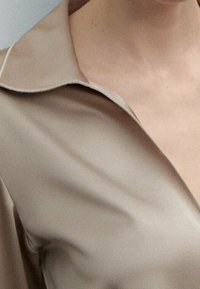Beżowa koszula z szerokim kołnierzem i gładką, miękką teksturą. Tkanina ma delikatny połysk, podkreślający jej elegancki, nowoczesny design.