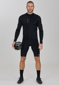 Schwarzes Radtrikot mit langen Ärmeln, Reißverschluss vorne und Logo. Enganliegende schwarze Shorts und schwarze Fahrradschuhe, während er einen schwarzen Helm hält.