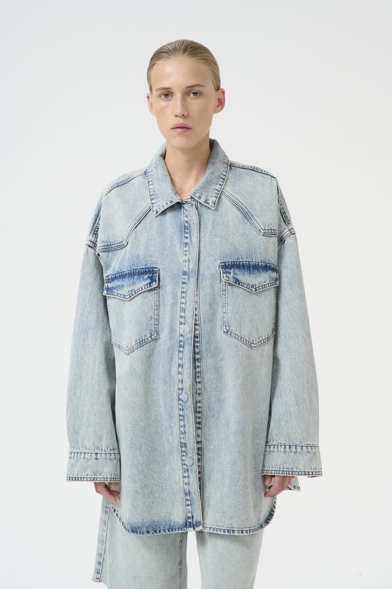 Giacca camicia oversized in denim azzurro chiaro con due tasche frontali, colletto appuntito e dettagli di cucitura a vista. Texture liscia.