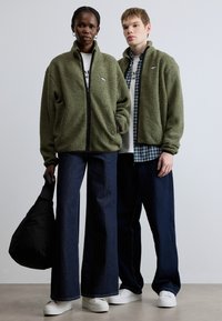 Grønne fleece-jakker med lynlås og tekstureret stof, kombineret med mørke, vidde jeans og hvide sneakers, mod en neutral baggrund.