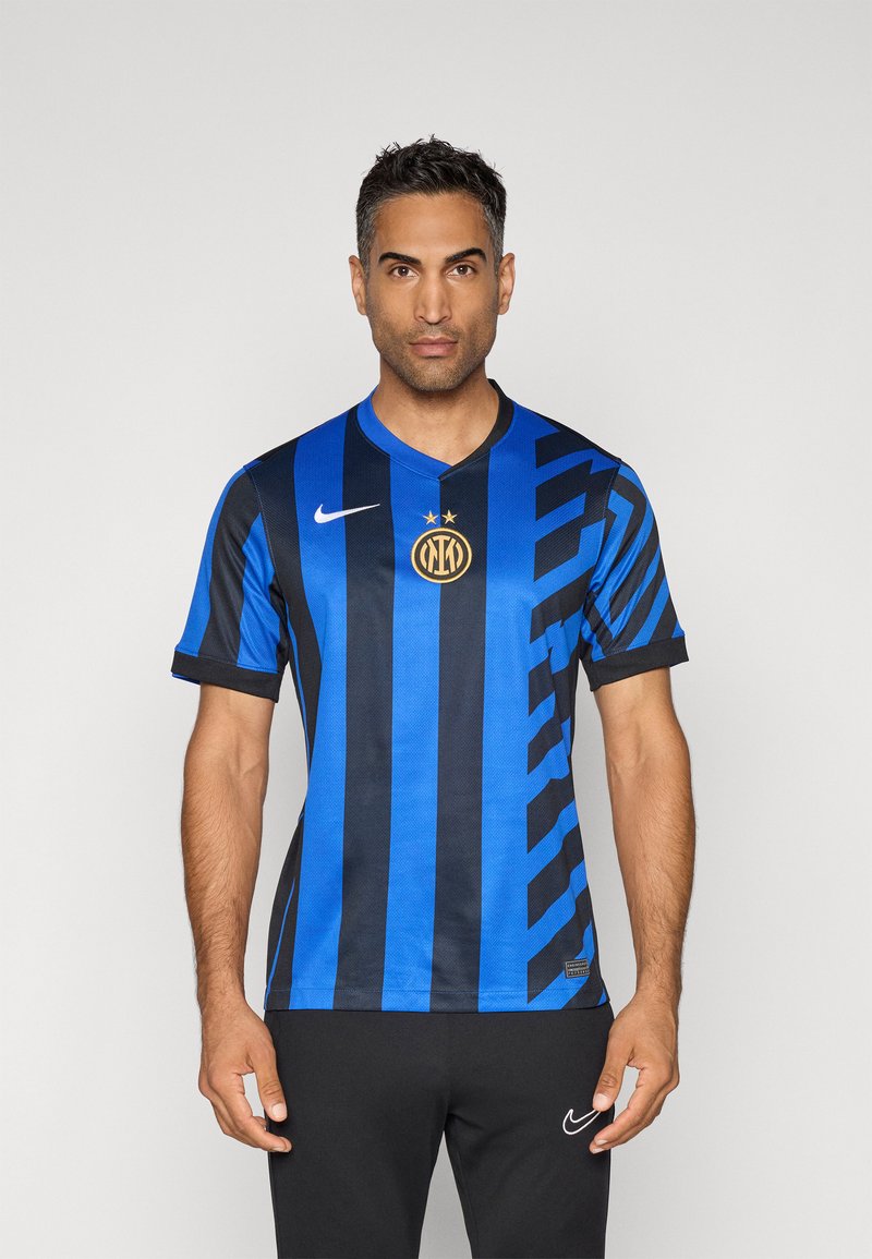 Hombre de pie vestido con una camiseta de fútbol de rayas azules y negras del Inter de Milán y pantalones negros de Nike, frente a un fondo claro y liso.