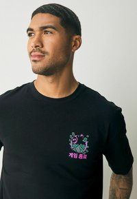 Tricou negru din bumbac, cu un grafic colorat al unui personaj de desene animate înconjurat de bani, având text în coreeană de culoare roz. Design cu mâneci scurte.