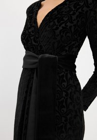 Schwarzes Samtkleid mit langen Ärmeln, verziert mit einem floralen Muster, tiefem V-Ausschnitt und einem breiten Satinträger, der in der Taille gebunden ist. Texturiertes Stoffdetail.
