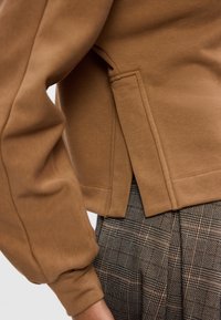 Personne portant un haut marron à manches longues avec une fente latérale et un pantalon à carreaux taille haute dans des tons noir et beige.