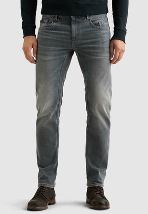 Homme portant un jean slim gris délavé, un pull noir et des bottes marron foncé à lacets, debout devant un fond uni.