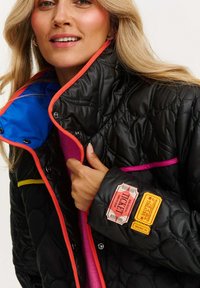 Prešívaná čierna bunda s jasne modrou podšívkou, oranžovými, ružovými a žltými akcentmi. Obsahuje jedinečné nášivky v štýle lístkov na rukávoch.