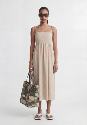 Mujer con vestido a cuadros beige y gafas de sol, sosteniendo un gran bolso tote de camuflaje, de pie frente a un fondo claro y liso.