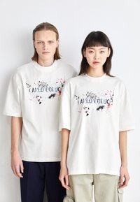 Krämfarvede oversized t-shirts med grafiske designs, der viser sprøjt af rosa, blå og sort, præsenteret af to modeller.