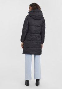 Manteau rembourré noir avec capuche, comportant des sections matelassées horizontales. Porté avec un jean large bleu clair et des bottes à talons noires.