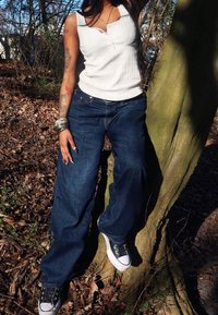 Person med tatueringar som bär en vit ärmlös topp, lösa blå jeans, svarta sneakers och silversmycken står mot ett träd i en höstskog.