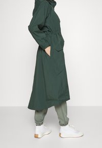 Zelený, lehký, dlouhý kabát se stahovacími šňůrkami v pase, dvěma bočními kapsami a kapucí, doplněný šedými tepláky a bílými teniskami.