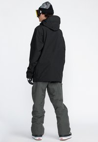 Veste imperméable noire avec une capuche, associée à un pantalon de neige gris ample. Présente une coupe décontractée et des gants à motifs.