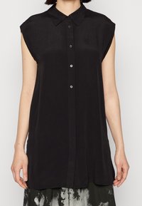 Kvinna klädd i en ärmlös svart knapp-up blus med spetsig krage, i kombination med en svart-vit mönstrad kjol.