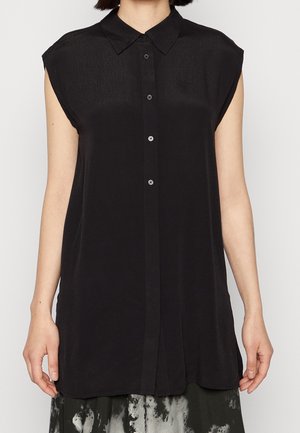 Femme portant un chemisier sans manches noir à boutons, avec un col pointu, associé à une jupe à motifs noir et blanc.