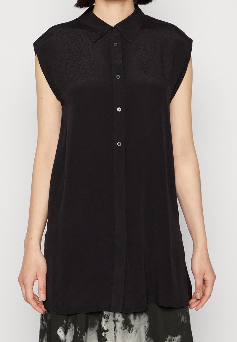 Kvinna klädd i en ärmlös svart knapp-up blus med spetsig krage, i kombination med en svart-vit mönstrad kjol.