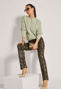 Frau trägt einen hellgrünen Kabelstrickpullover, Schlangenprint-Hose, beigefarbene High Heels, hält eine metallische Clutch und eine runde Sonnenbrille.