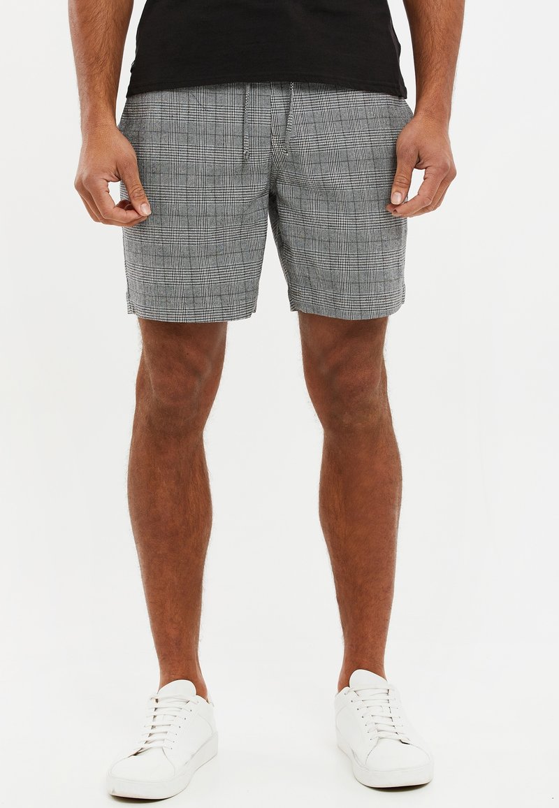 Homme portant un short gris à carreaux, un t-shirt noir et des baskets blanches, debout devant un fond blanc uni.