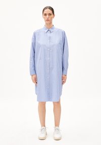 Robe chemise à boutons, rayée bleu et blanche, avec un col et des manches longues. Tissu doux, longueur genoux, associée à des baskets blanches.