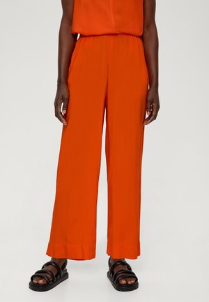 Person iført klare orange bukser med vide ben og sorte sandaletter med remme, stående mod en ensfarvet lys baggrund.