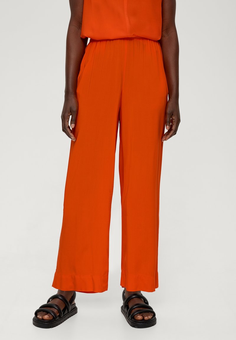Personne portant un pantalon large orange vif et des sandales noires à brides, debout devant un fond uni clair.
