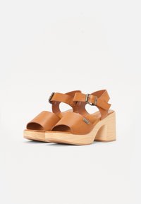 Sandálias de couro castanho com um design de biqueira aberta, tiras ajustáveis para o tornozelo e saltos de madeira chunky. Apresenta um detalhe em metal com o logótipo.