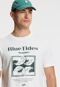 Balta medvilninė marškinėliai su žalia bangų grafika ir užrašu "BlueTides Resort." Taip pat rodomi žali kepuraitė su mažu logo.