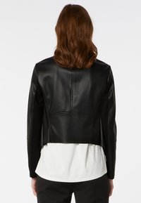 Černá kožená zkrácená bundička s hladkou texturou, s vystouplými rameny a čistým designem. Nositá přes bílou košili.