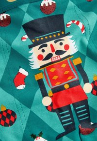 Bomuldsstof med en nøddeknækkerkarakter i en rød og sort uniform mod en teal baggrund med festlige detaljer som sukkerrør og ornamenter.