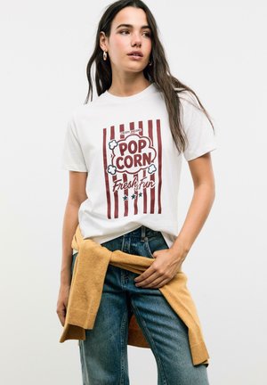 T-shirt blanc avec un motif de popcorn rouge et blanc accompagné de texte. Porté avec un jean bleu et un pull marron clair noué à la taille.