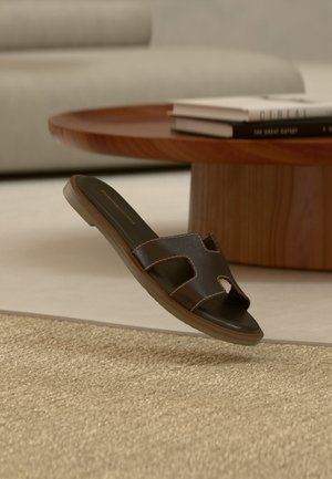 Sandalia deslizable de cuero marrón oscuro sobre alfombra beige, con mesa de café redonda de madera y libros apilados en el fondo.