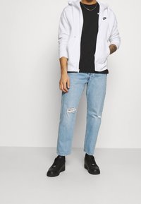 Man som bär en vit Nike-hoodie, svart Nike-T-shirt, ljusblå slitna jeans, svarta sneakers och guldhalsband framför en platt vit bakgrund.