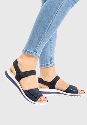 Füße in marineblauen offenen Sandalen mit Klettverschlüssen und weißen Sohlen, kombiniert mit hellblauen Skinny-Jeans vor einem einfachen Hintergrund.