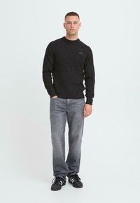 Sort striktrøje med rund hals og tekstureret mønster, parret med lysegrå jeans og sorte sneakers. Modellen står i en neutral stilling.
