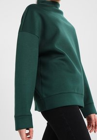 Persoon in een donkergroene sweatshirt met hoge kraag, lange mouwen en zwarte broek tegen een effen grijze achtergrond.