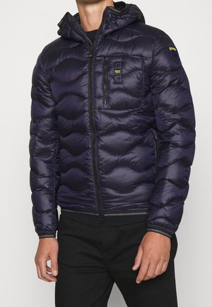 Mann trägt eine dunkelblaue gesteppte Daunenjacke mit Kapuze, Frontreißverschluss, Brusttasche und schwarze Hose.